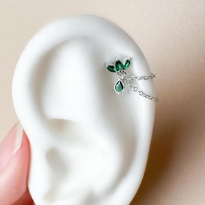 May include: A silver ear cuff featuring a cluster of green gemstones and a dangling teardrop-shaped gem. The cuff is connected by a delicate chain. The jewelry is displayed on a white ear model.