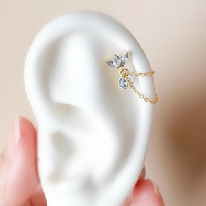 May include: A gold-coloured ear piercing with a floral design, three light blue gemstones, and a dangling teardrop-shaped gemstone. A delicate chain connects the piercing to the ear. The jewellery is displayed on a white ear model.