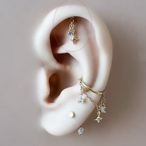 May include: Gold-toned ear piercings with dangling charms. The top piercing features a chain with a blue gem and a star charm. Additional piercings include a hoop, studs, and dangling charms with gems and stars. The ear is set against a neutral background.
