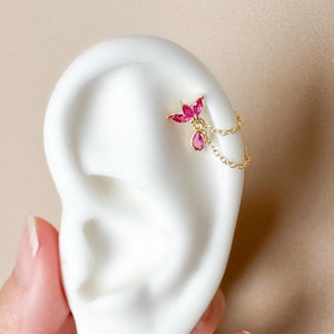 May include: A gold-colored ear piercing featuring a cluster of pink gemstones in a floral design, connected by a delicate chain to a dangling teardrop-shaped pink gemstone. The jewelry is displayed on a white ear model against a neutral background.
