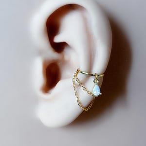 May include: A gold-colored ear cuff featuring a delicate chain and a teardrop-shaped, iridescent gemstone. The cuff is designed to wrap around the ear's cartilage, adding a touch of elegance to any look. The jewelry is set against a neutral background.