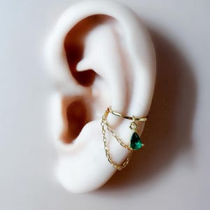 May include: Gold-colored ear cuff with a dangling emerald-green teardrop-shaped gem and delicate chain accents. The cuff wraps around the ear, adding a touch of elegance. A small, clear gem is also present.