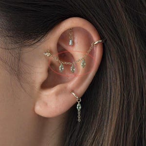 May include: Gold-coloured ear jewellery featuring a chain with three dangling green gemstones. A small hoop earring with a dangling green gemstone is visible. Another piece of jewellery is above the chain.