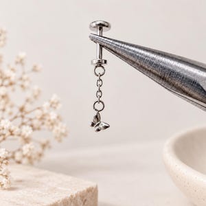 May include: A silver-toned metal body jewelry piece featuring a flat circular backing, a straight post, and a delicate dangling chain attachment that ends in a small, detailed butterfly charm. The jewelry is held by metal pliers.