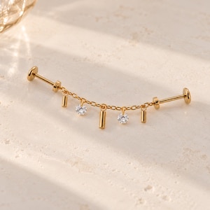 May include: A gold-toned industrial barbell piercing jewelry piece featuring a central chain draped between two posts. The chain is adorned with three small vertical gold bars and two round clear faceted stones hanging as charms.