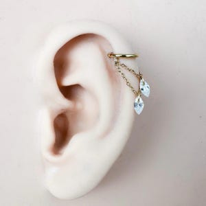 May include: A gold-colored ear cuff with a dangling chain and two clear, teardrop-shaped gemstones. The cuff is attached to the ear, showcasing the jewelry's design and placement. The gemstones add a touch of sparkle.
