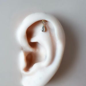 Puede incluir: Un pendiente de color dorado con una pequeña gema azul claro en forma de lágrima cuelga de la oreja. El pendiente está sujeto a la oreja con un pequeño perno redondo. La oreja es de tono de piel claro y el fondo es de color neutro.