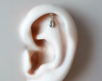May include: A gold-colored earring with a small, teardrop-shaped, light blue gemstone hangs from the ear. The earring is attached to the ear with a small, round stud. The ear is a light skin tone, and the background is a neutral color.