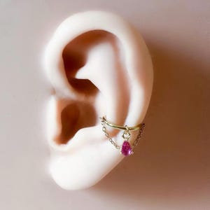 May include: A gold-colored ear cuff with a small chain and a dangling pink teardrop-shaped gemstone. The cuff is attached to a model ear, showcasing the jewelry's design and placement.