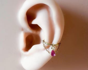 May include: A gold-colored ear cuff with a small chain and a dangling pink teardrop-shaped gemstone. The cuff is attached to a model ear, showcasing the jewelry's design and placement.