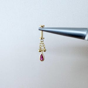 以下が含まれることがあります： ぶら下がりデザインのゴールドイヤリング。イヤリングは、下部に小さな涙型の赤い宝石があり、その上に小さなゴールドチェーンとビーズが連なっています。イヤリングは、白い背景に対して銀色のツールで保持されています。