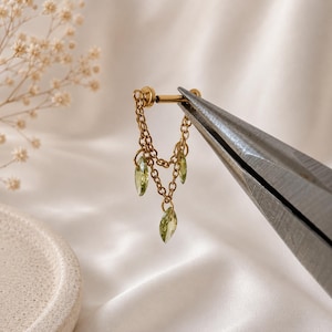 Op de afbeelding: Een goudkleurige metalen oorbel met een rechte stift en twee fijne kettinkjes die naar beneden hangen. Drie kleine, lichtgroene, doorschijnende steentjes in marquise-slijpvorm zijn aan de uiteinden van de kettinkjes bevestigd voor een gelaagd effect.