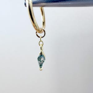 May include: A gold hoop earring with a small, teardrop-shaped pendant. The pendant features a light blue and green stone with a unique, natural pattern. The earring is suspended from a metal bar, showcasing the jewelry's design.