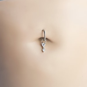 Puede incluir: Un piercing para el ombligo plateado con dos piedras de circonita cúbica transparentes y brillantes. La piedra superior es redonda y la inferior tiene forma de corazón. El anillo está sujeto a una barra curva.