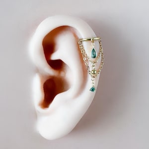 May include: Gold-toned ear cuff with dangling chains and teal teardrop-shaped gems. The ear cuff is attached to a model ear, showcasing the jewelry's design and placement. The design includes a small gold ball and multiple chains.