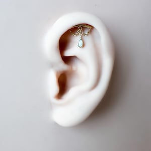 May include: A gold-colored ear piercing with a chain and a teardrop-shaped, opalescent charm. The jewelry is set against a light background, highlighting the intricate design and delicate details of the earring.