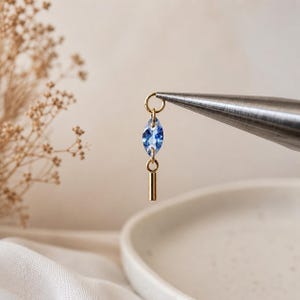 May include: A gold-toned jewelry charm featuring a marquise-cut blue crystal suspended from a small circular jump ring, with a short, smooth gold-toned cylindrical bar dangling vertically from the bottom of the crystal, held by metal pliers.