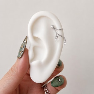 May include: A silver-toned cartilage earring featuring a small ball stud connected by two delicate chains to two clear, teardrop-shaped crystal dangles. The earring is displayed on a white ear model, showcasing a multi-chain design.