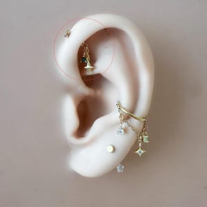 May include: Close-up of an ear adorned with multiple gold earrings. One earring features a dangling star charm with a small green gem. Other earrings include a small stud and a chain with dangling charms.