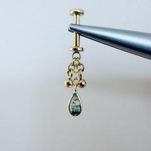 May include: A gold earring with a chain of small gold beads and a teardrop-shaped pendant. The pendant has a green and white pattern. The earring is held by a silver tool.