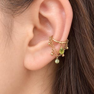 May include: A gold-toned ear cuff featuring multiple delicate chains draped across the cartilage. The design includes small gold spheres and two light green, teardrop-shaped faceted stones suspended from the chains, creating a layered effect.