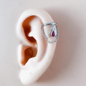 May include: A silver ear cuff with a dangling chain design. The cuff features a small, teardrop-shaped, red gemstone pendant. The jewelry is designed to be worn on the ear cartilage.