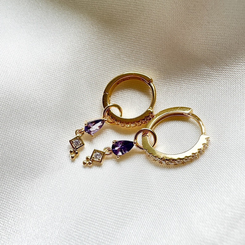 May include: Gold hoop earrings with a dangling purple teardrop and clear crystal accents.