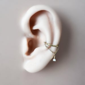 May include: A gold ear cuff featuring a fine chain and a small, pear-shaped, white gemstone. The cuff wraps around the ear, with a chain that hangs down. The jewellery is displayed on a light-coloured ear.