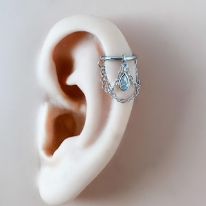 May include: Silver ear cuff featuring a dangling chain design with a teardrop-shaped clear gemstone. The cuff is attached to a model ear, showcasing the jewelry's placement and style. The ear cuff is a delicate piece of body jewelry.