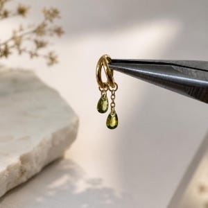 May include: A gold hoop earring featuring two dangling charms in the shape of teardrops. Each charm is set with a small, faceted olive green gemstone. The earring is held by a pair of metal pliers, showcasing the delicate chain and metal links.