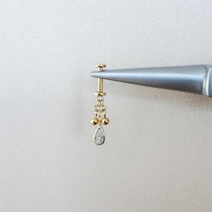 May include: A gold-colored earring with a dangling design, held by a silver tool. The earring features a small, pear-shaped clear stone at the bottom, with gold beads and chains above.