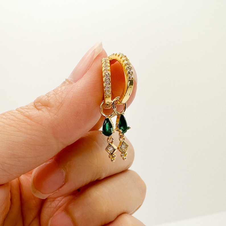 May include: Gold hoop earrings with a dangling emerald green teardrop charm and a small diamond-shaped charm with a row of small gold beads.
