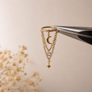 May include: A gold-toned hoop earring featuring three delicate dangling chains. The center chain holds a small crescent moon charm, while the outer chains support a clear round gemstone and a tiny four-pointed star charm at the base.