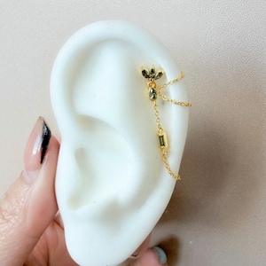 May include: Gold-colored ear jewelry featuring a floral design with small, dark gemstones and a dangling chain with a rectangular gem. The jewelry is attached to a white ear model.