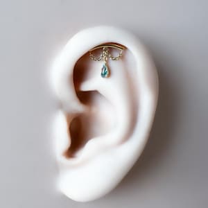 May include: A gold-colored ear piercing featuring a curved bar with dangling chains and a teardrop-shaped, teal-colored gemstone. The jewelry is displayed on a white ear model, showcasing the design and details of the piercing.