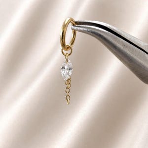 Puede incluir: Un pendiente de aro dorado con un dije de piedra transparente en talla marquesa suspendido de una cadena dorada corta y delicada. El aro está sujeto por unos alicates de metal, mostrando el acabado dorado pulido y la piedra facetada.