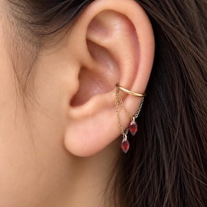 May include: A gold ear cuff featuring two delicate chains that drape downward, each ending in a small, faceted, marquise-cut deep red gemstone charm. The smooth, circular gold band secures the piece to the upper cartilage of the ear.