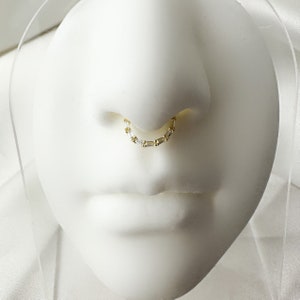 Può includere: Un anello per il naso in oro con una catena di cristalli trasparenti. L'anello è su una testa di manichino bianca.