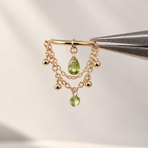 Könnte beinhalten: Ein goldfarbener Ohrring mit zwei zarten, drapierten Ketten. Das Design umfasst kleine goldene Kugeln an den Ketten, einen zentralen birnenförmigen hellgrünen Edelstein und einen runden hellgrünen Edelstein, der in der Mitte unten hängt.