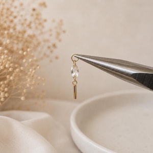 May include: A gold-toned earring component featuring a small, clear, marquise-cut crystal bead suspended from a jump ring, with a slender, polished gold-toned cylindrical bar dangling at the bottom, held by a pair of metal pliers.