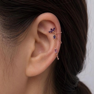 May include: Gold-toned ear cuff featuring a cluster of purple gemstones at the top, with dangling chains and additional purple gems. The jewelry is worn on the ear, showcasing the design and color.