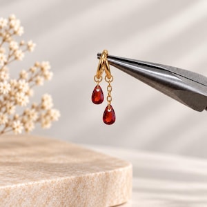 May include: A gold hoop earring featuring two dangling red teardrop-shaped crystals of different lengths. One crystal hangs directly from the hoop, while the other is suspended by a short gold chain, held by metal pliers.