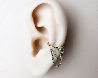 Peut inclure: Manchette d'oreille dorée ornée d'une petite pierre précieuse bleu clair et de chaînes pendantes. Le bijou est conçu pour s'enrouler autour du cartilage de l'oreille. La manchette est présentée sur un modèle d'oreille de couleur chair pâle.