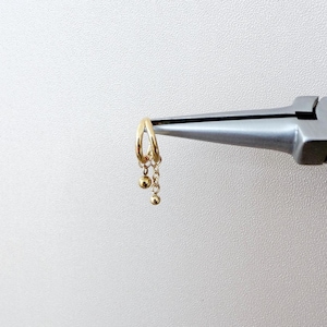 May include: A gold-colored hoop earring with dangling ball charms. The earring is held by a silver tool against a white background. The earring has a simple, elegant design.