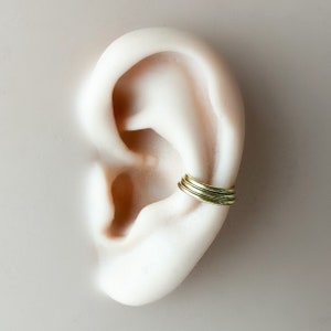 May include: A gold ear cuff with multiple rings stacked on top of each other. The cuff is worn on the upper part of the ear, just above the earlobe.