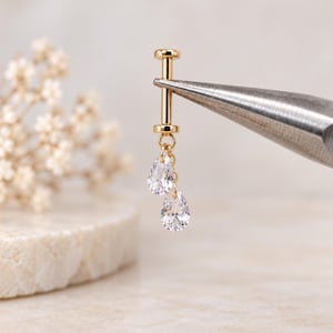 May include: A gold-toned flat back labret stud featuring a vertical post with two dangling clear, pear-shaped cubic zirconia charms. The jewelry piece is held by metal pliers, showcasing the polished gold finish and the faceted, teardrop-shaped stones.