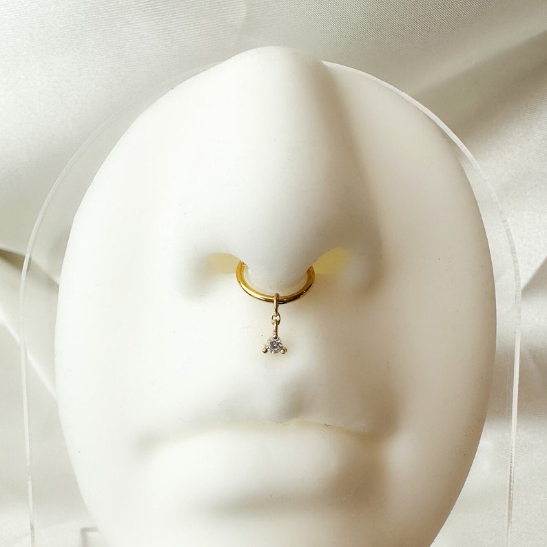 K&ouml;nnte beinhalten: Ein goldener Nasenring mit einem kleinen, klaren Edelstein, der daran h&auml;ngt.