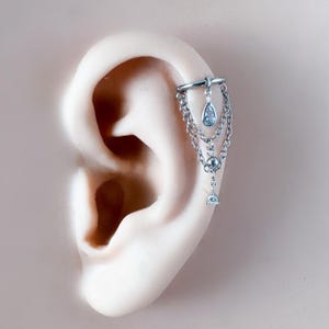 May include: Silver ear cuff with dangling chains and teardrop and round-cut crystal accents. The ear cuff is attached to the ear with a small hoop and features multiple chains with crystal embellishments.