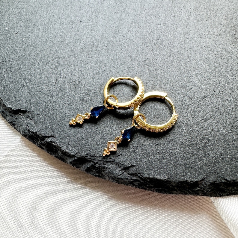 May include: A pair of gold hoop earrings with a dangling blue gemstone and clear crystal accents.