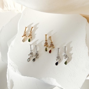 May include: An assortment of gold and silver-toned earrings with dangling teardrop-shaped gemstones. The earrings feature various colored gems, including clear, green, and red. The earrings are displayed on a white, textured surface.